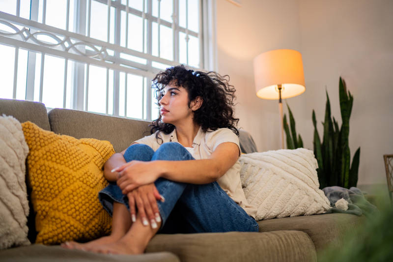 A woman sitting on a couch, looking out a window, feeling anxiety and stress.